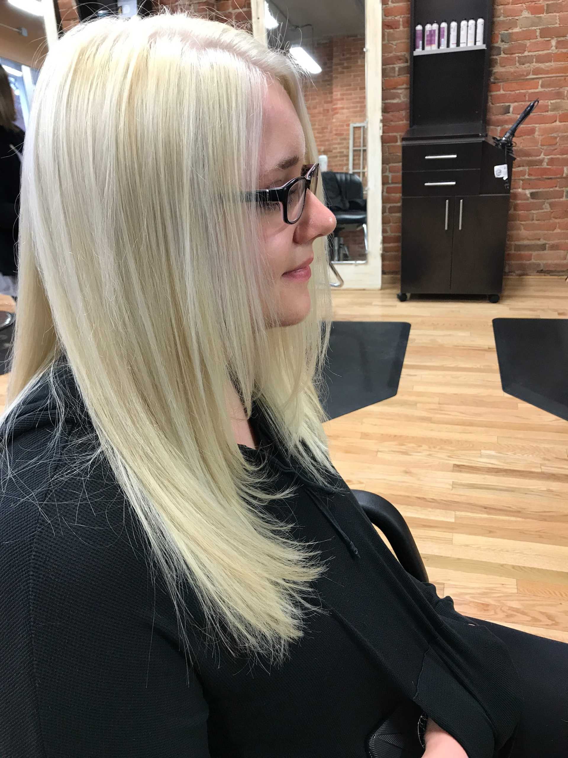 Person with long blonde hair and glasses sitting in a brick-walled salon.