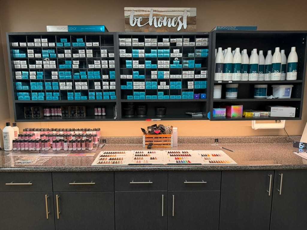 Hair salon color station with shelves of dye boxes and bottles, color charts on counter.