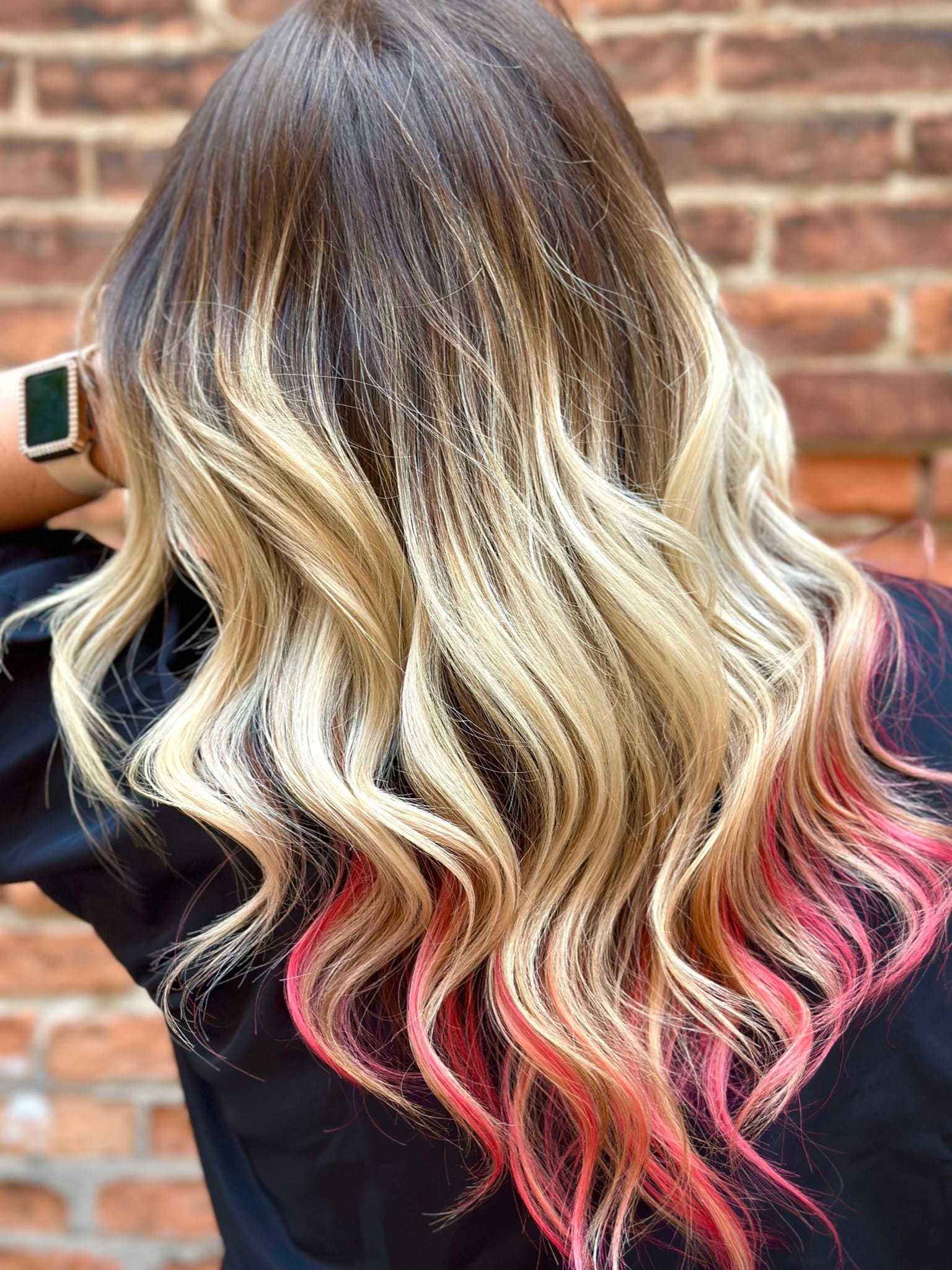 Long wavy hair with blonde highlights and pink tips against a brick wall background.