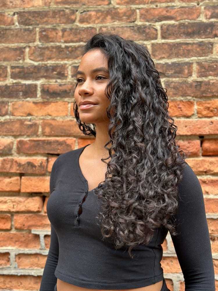 Woman with long curly hair, in a black top, standing against a brick wall.