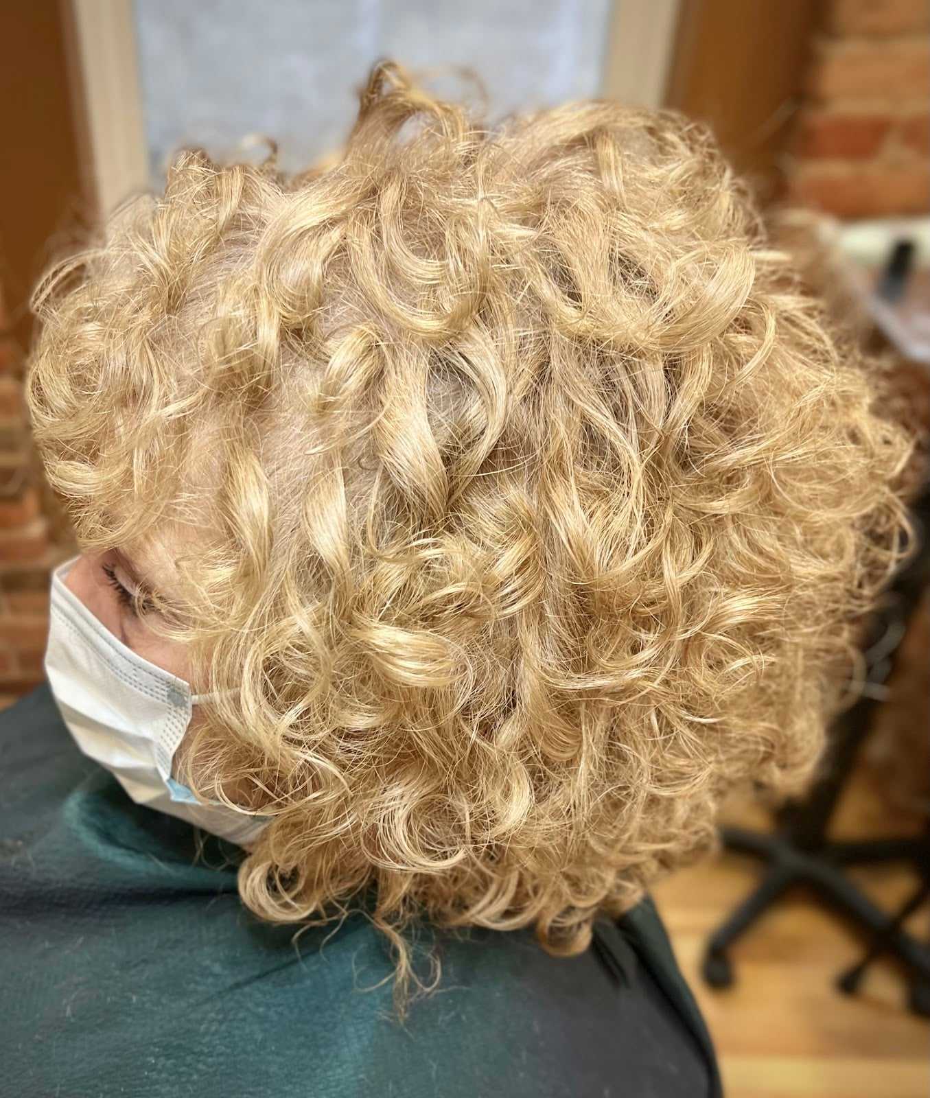 Curly blonde hair styled in a layered cut, person wearing a mask in a salon setting.