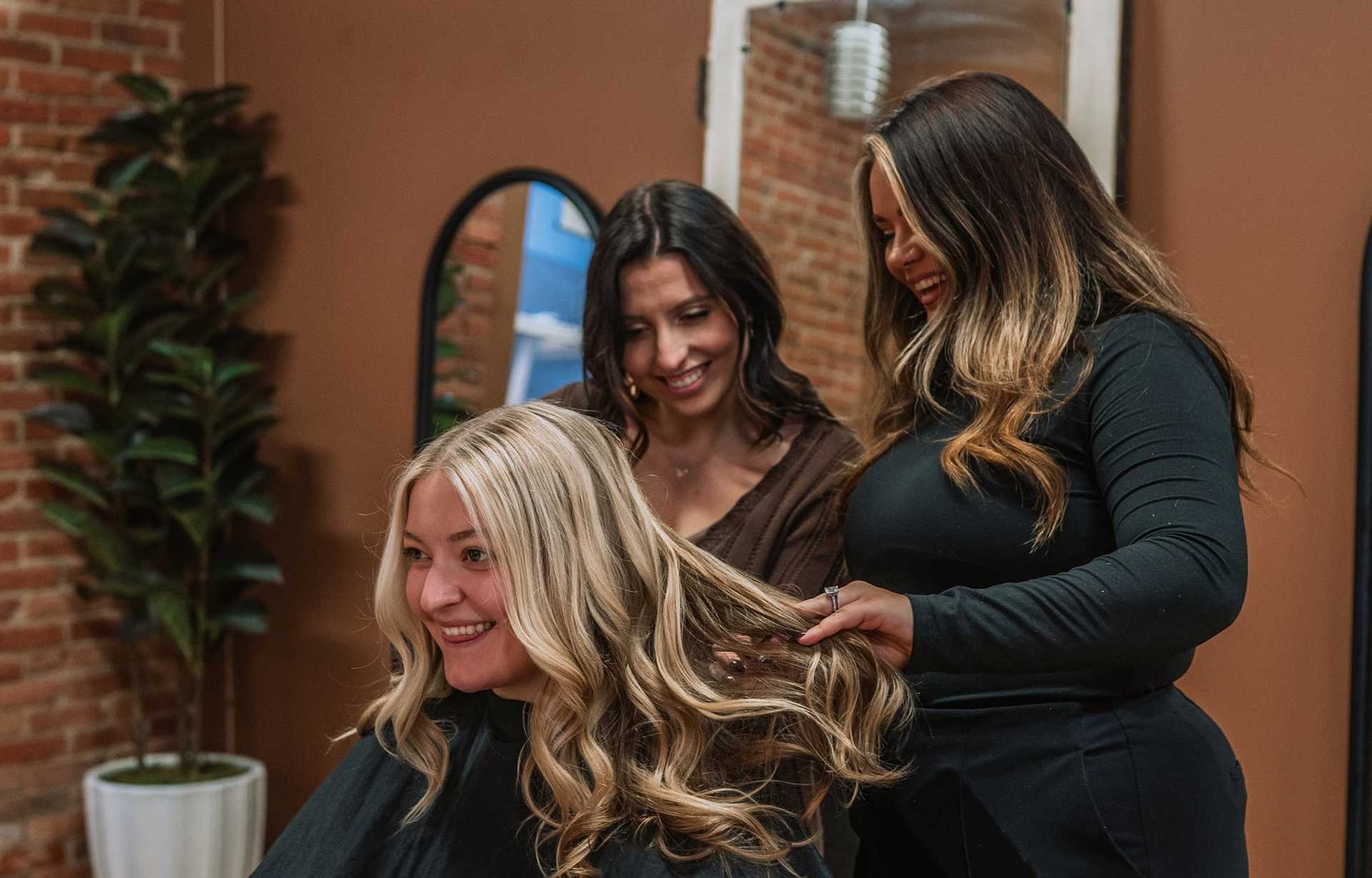 Three women in a salon smile as one styles another's long wavy blonde hair.