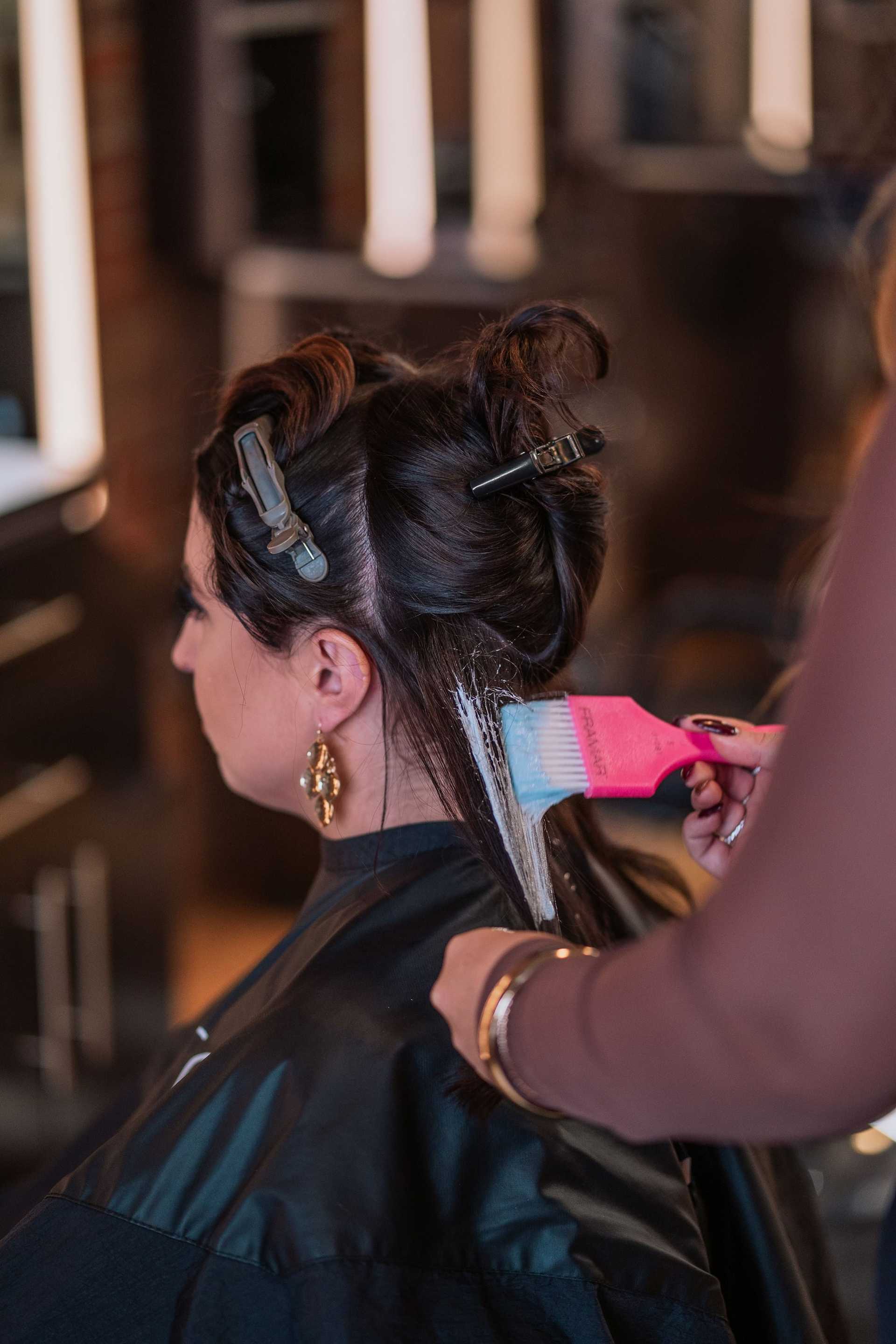 A hair stylist applies color to a woman's hair in a salon setting.