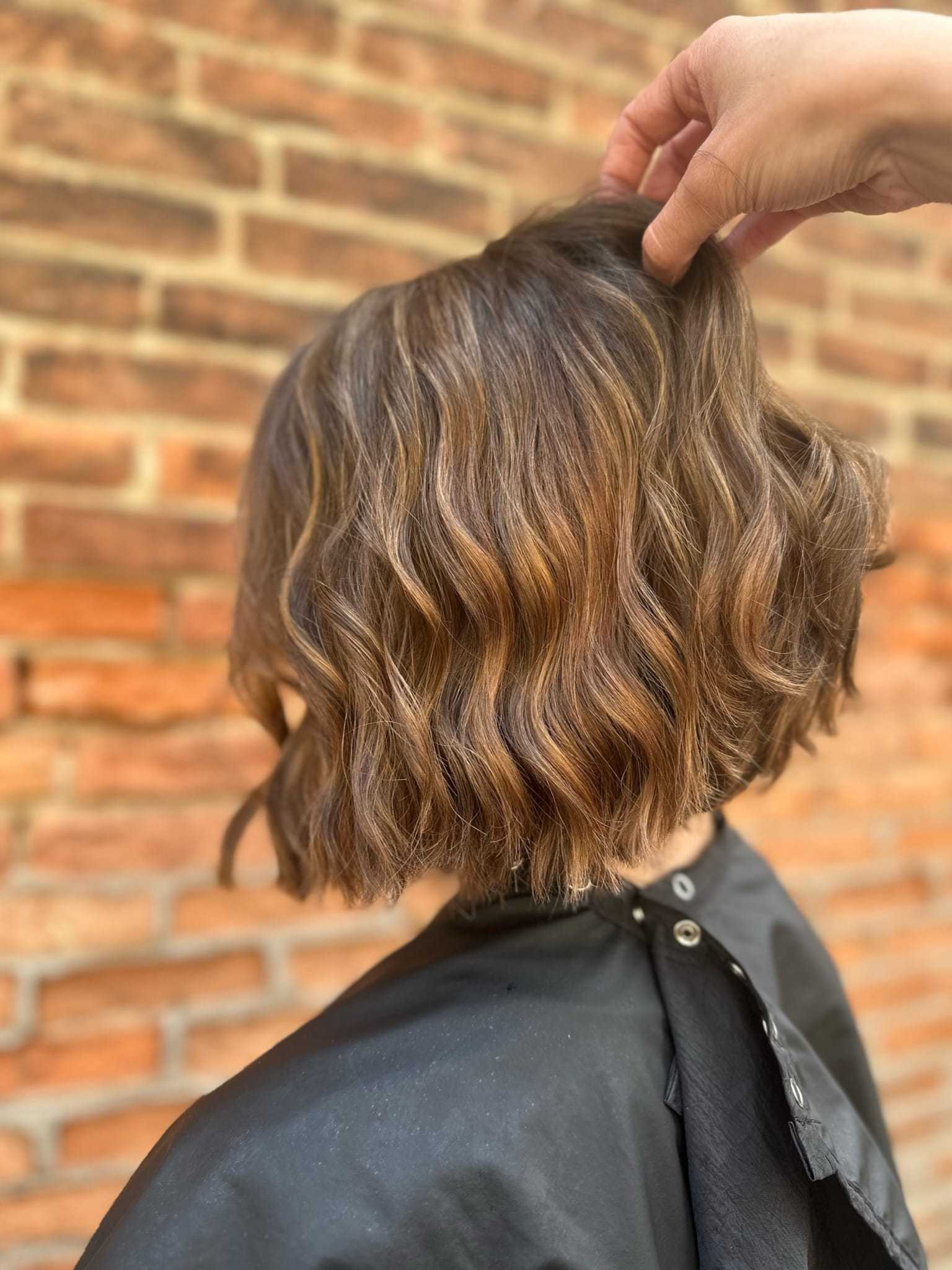 Wavy brown bob hairstyle with subtle highlights against a brick wall background.